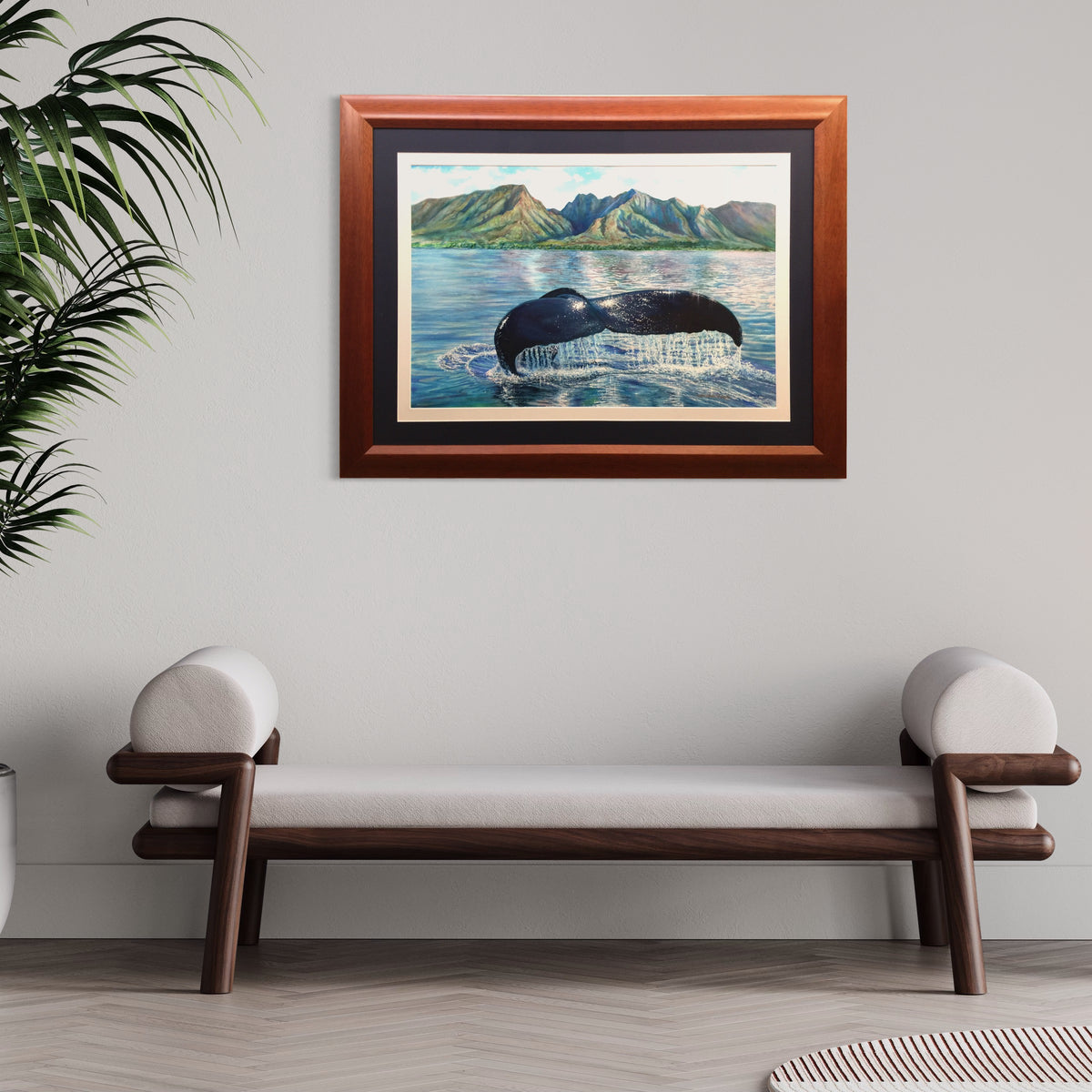 Professionally framed  watercolor art  depicting a humpback whale tail surfacing in Hawaiian blue water with green mountains in the background, hung on a light gray wall above a wooden bench with white cushions.